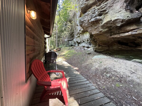 A wooden deck with a grill and red chairs, a rock wall, and a wooden cabin.