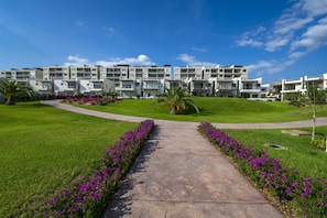 A modern apartment complex with a central pathway, surrounded by well-maintained lawns and vibrant flower beds. - NEW CONDO 3BR/3BTH with arch view at Vista Vela condos (Cabo San Lucas)
