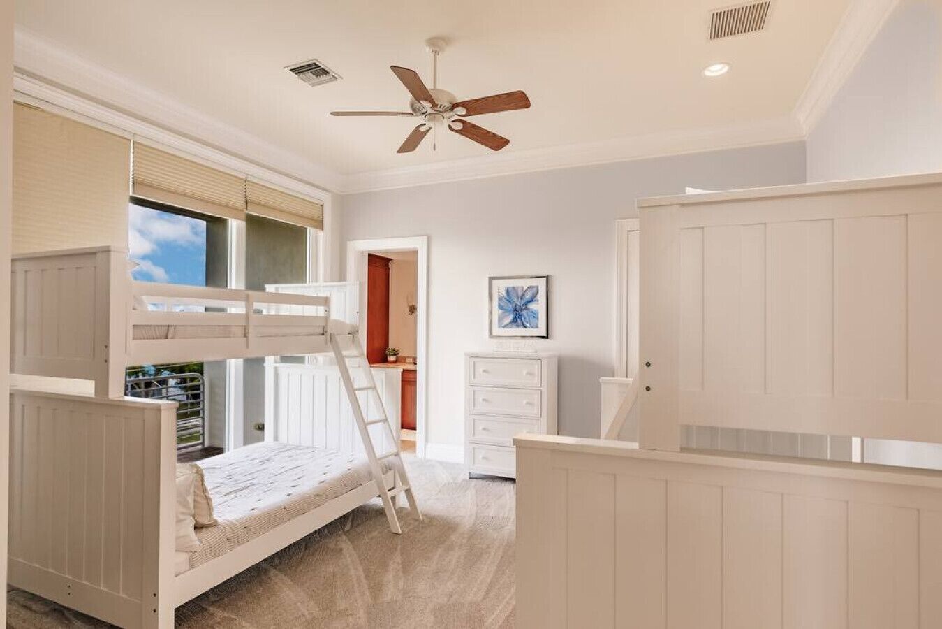 Une chambre avec un lit superposé, un ventilateur de plafond, une fenêtre avec des stores, une porte donnant sur la cuisine et un tableau encadré au mur.