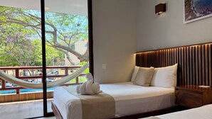 Una habitación de hotel con cama, mesita de noche, una hamaca y vistas a árboles y edificios.