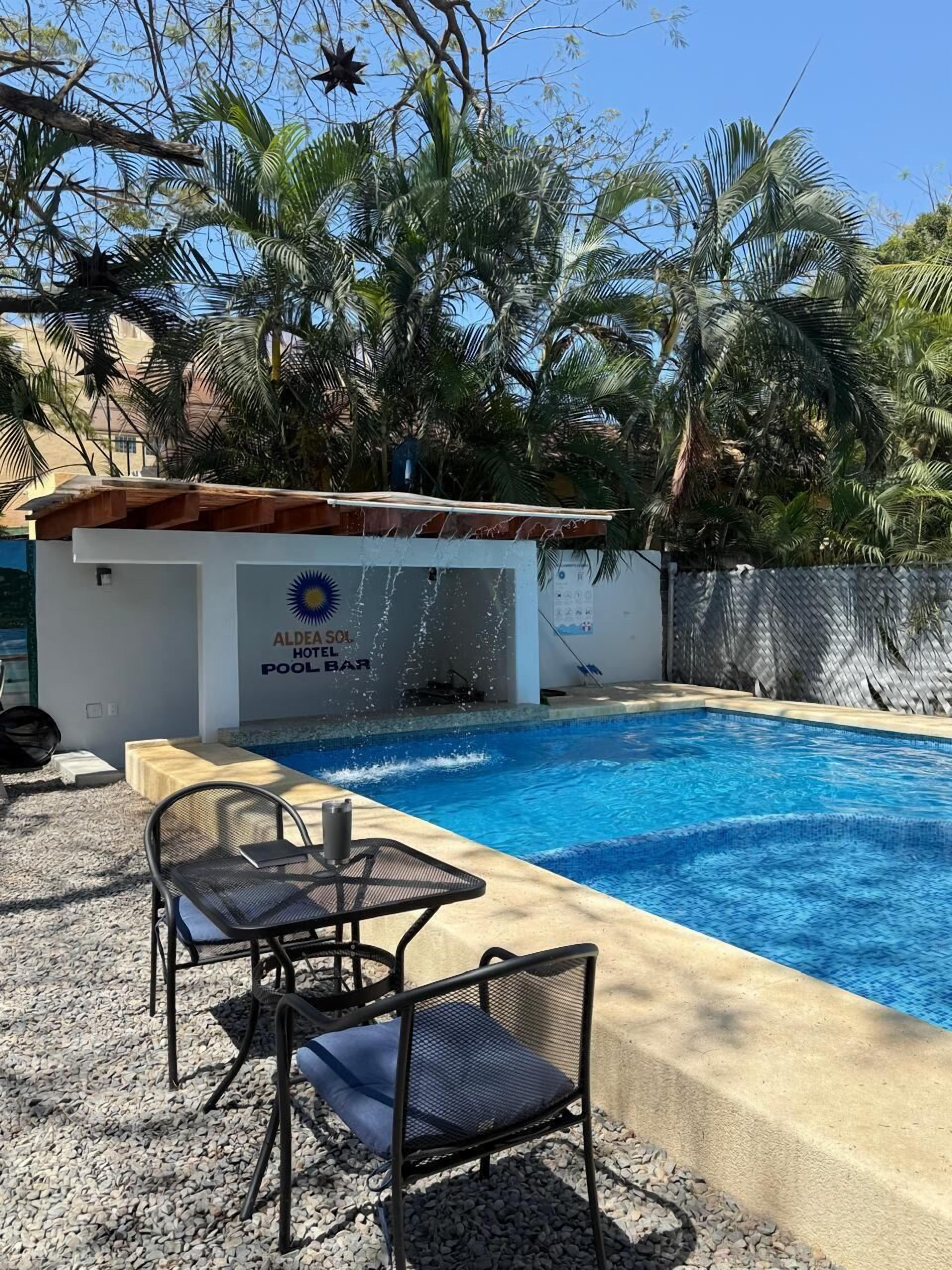 A swimming pool with a water feature, surrounded by palm trees and a building with a sign reading "Aldeia Sol Hotel Bar".