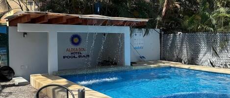 A swimming pool with a water feature, surrounded by palm trees and a building with a sign reading "Aldeia Sol Hotel Bar".