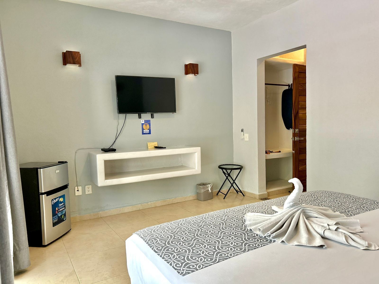 Una habitación de hotel con cama, televisor montado en la pared, minibar y una mesita.