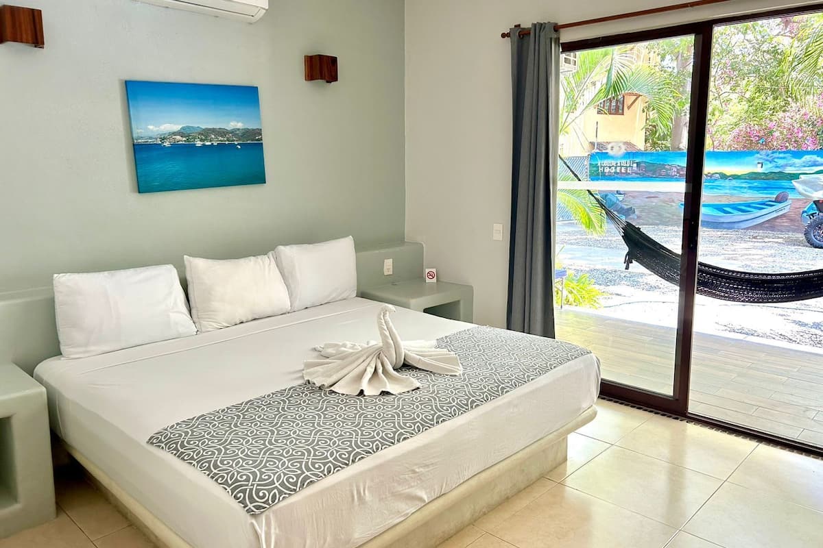 A hotel room with a bed, a nightstand, a wall-mounted air conditioner, a painting, and a view of boats and greenery outside.