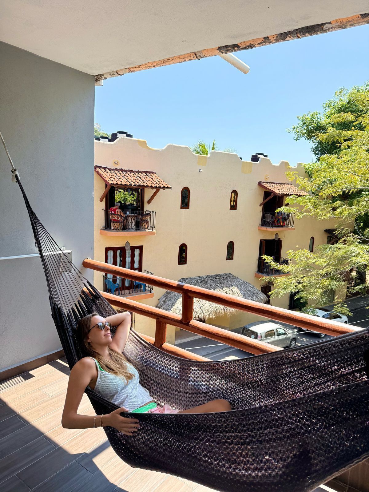 A person relaxing in a hammock on a balcony with a view of a building with balconies and a thatched roof structure.