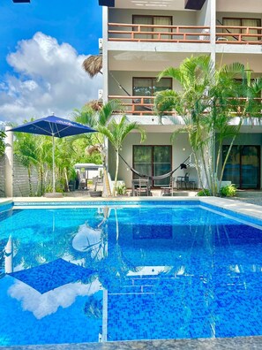 A swimming pool with a clear blue water, surrounded by a modern building with balconies and palm trees. - Hotel Aldea Sol (Zihuatanejo)