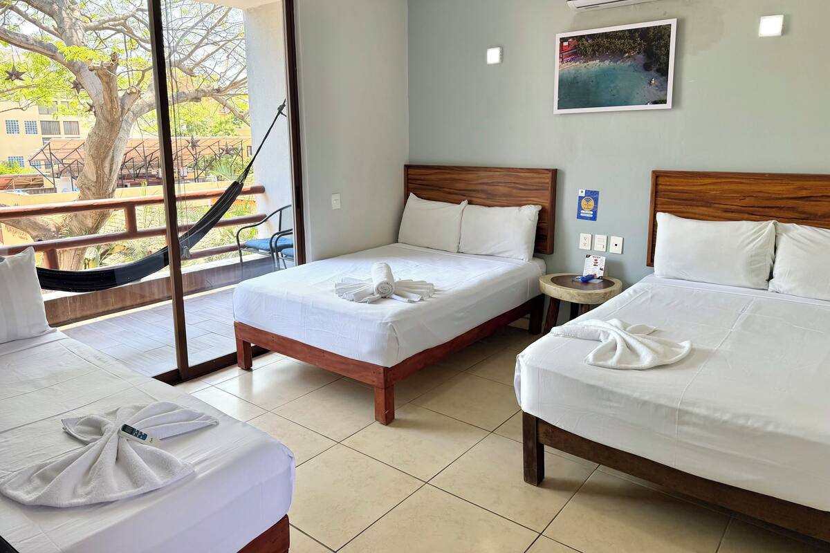 A hotel room with two beds, a small round table, a hammock, and a large window with a view of trees and buildings.