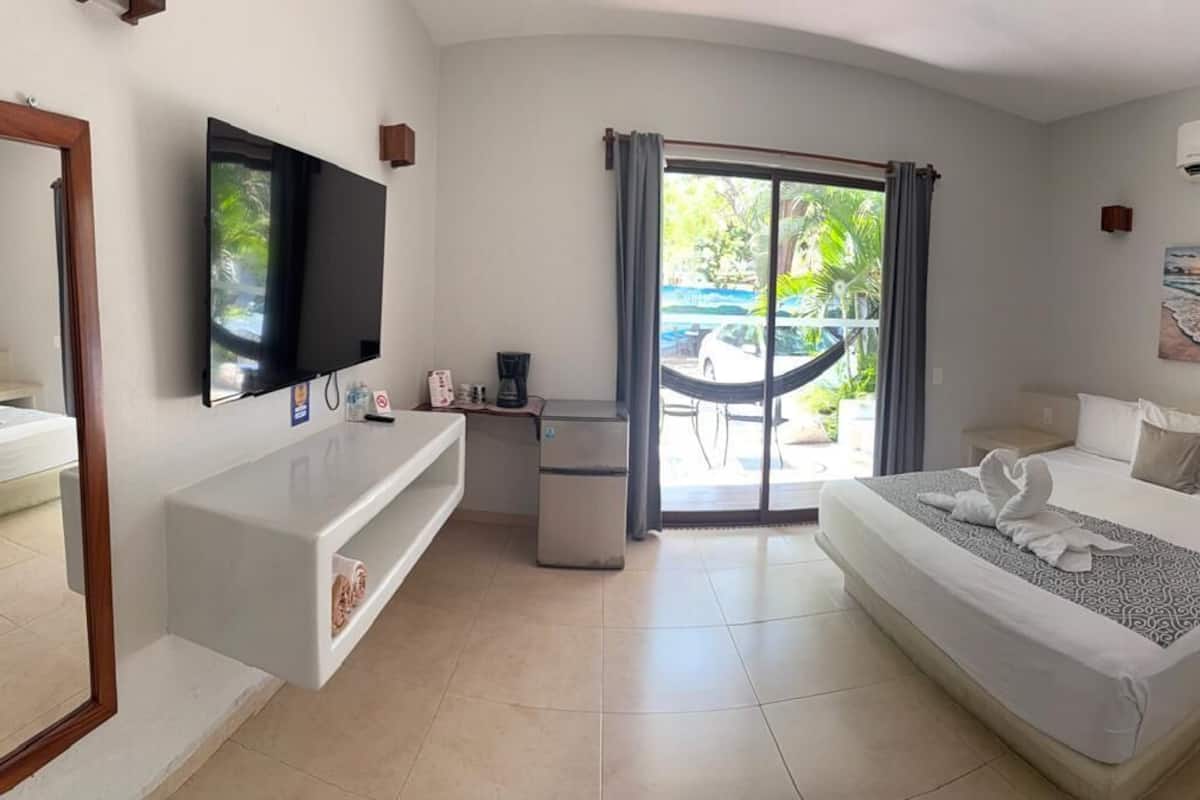 A hotel room with a bed, a TV, a desk, a chair, a small table, a trash bin, a sliding door to a balcony, and a painting on the wall.
