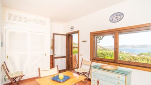Un coin repas avec une table en bois, des chaises, un set de table bleu et une vue sur la mer par la fenêtre.