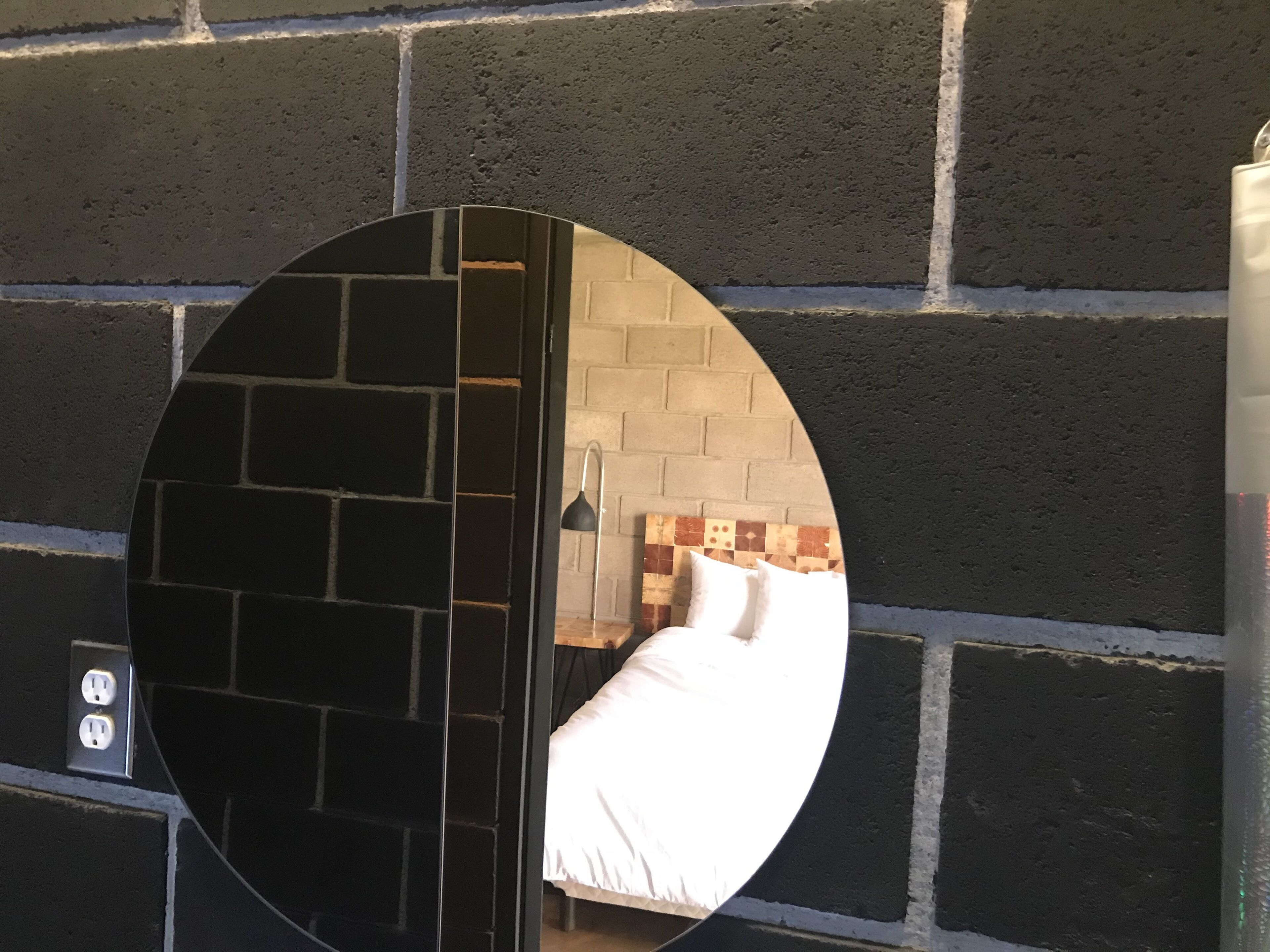 A circular mirror reflecting a room with a brick wall and a bed with white bedding.
