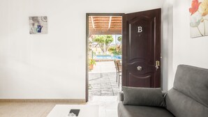 Een kamer met een houten deur met de aanduiding 'B', een grijze bank, een witte salontafel en uitzicht op een terras met een zwembad en planten.