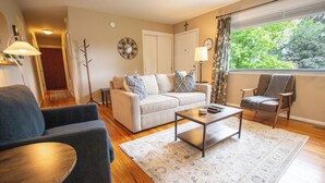 Ein Wohnzimmer mit einer Couch, Sesseln, einem Couchtisch und Blick auf Bäume durch ein Fenster.