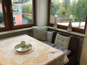 Un rincón con asientos, una mesa, cojines y una ventana que ofrece vistas a edificios y árboles.