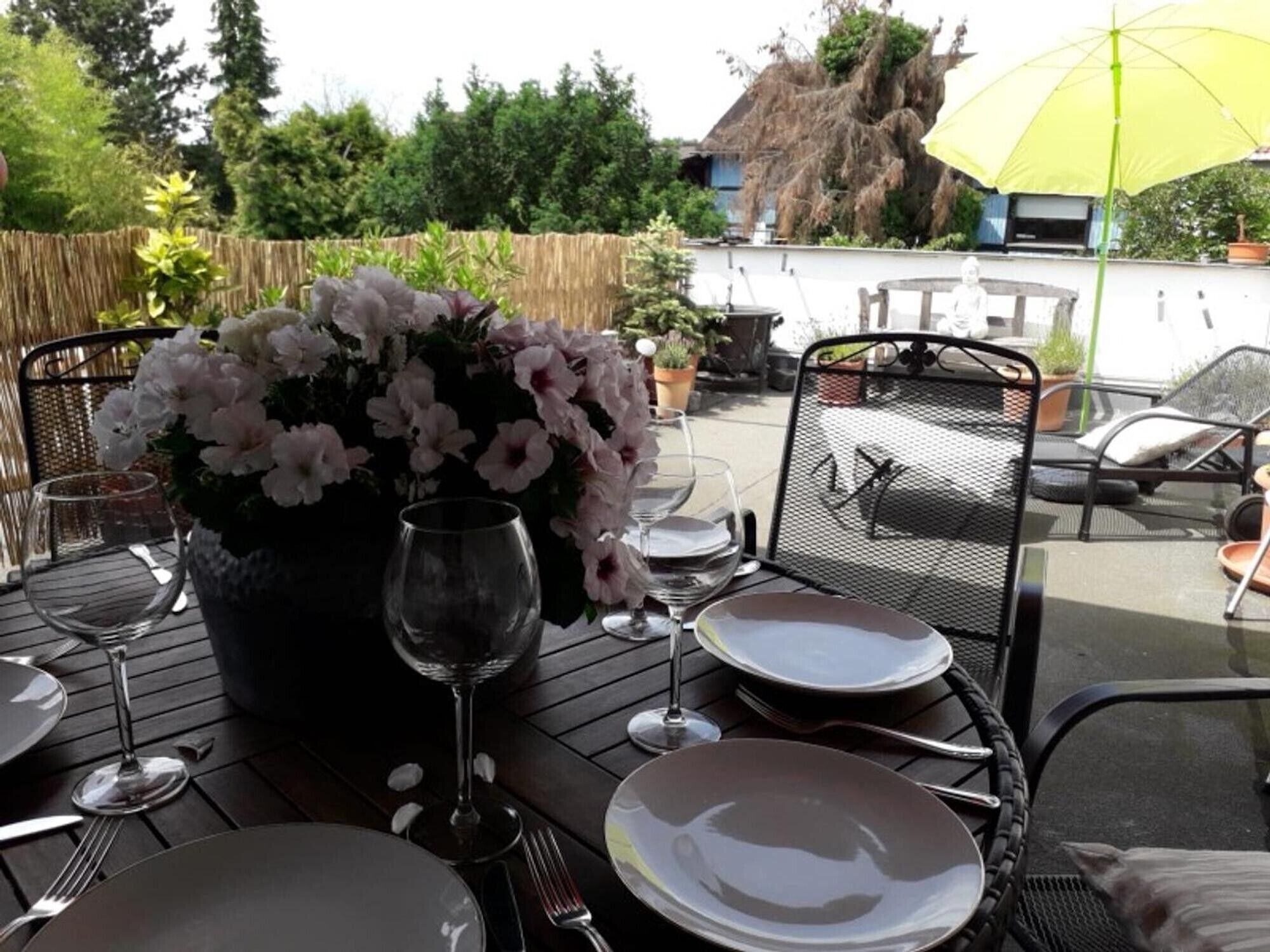 Un'area pranzo all'aperto con un tavolo apparecchiato per due persone, una pianta in vaso con fiori e un ombrello giallo.