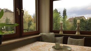 A room with a table set for two, a window with a view of trees and buildings, and a potted plant on the windowsill.