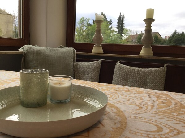 Ein Tisch für zwei ist gedeckt, ein Fenster bietet Blick auf Bäume und Gebäude, und zwei Kerzen stehen auf dem Fensterbrett.