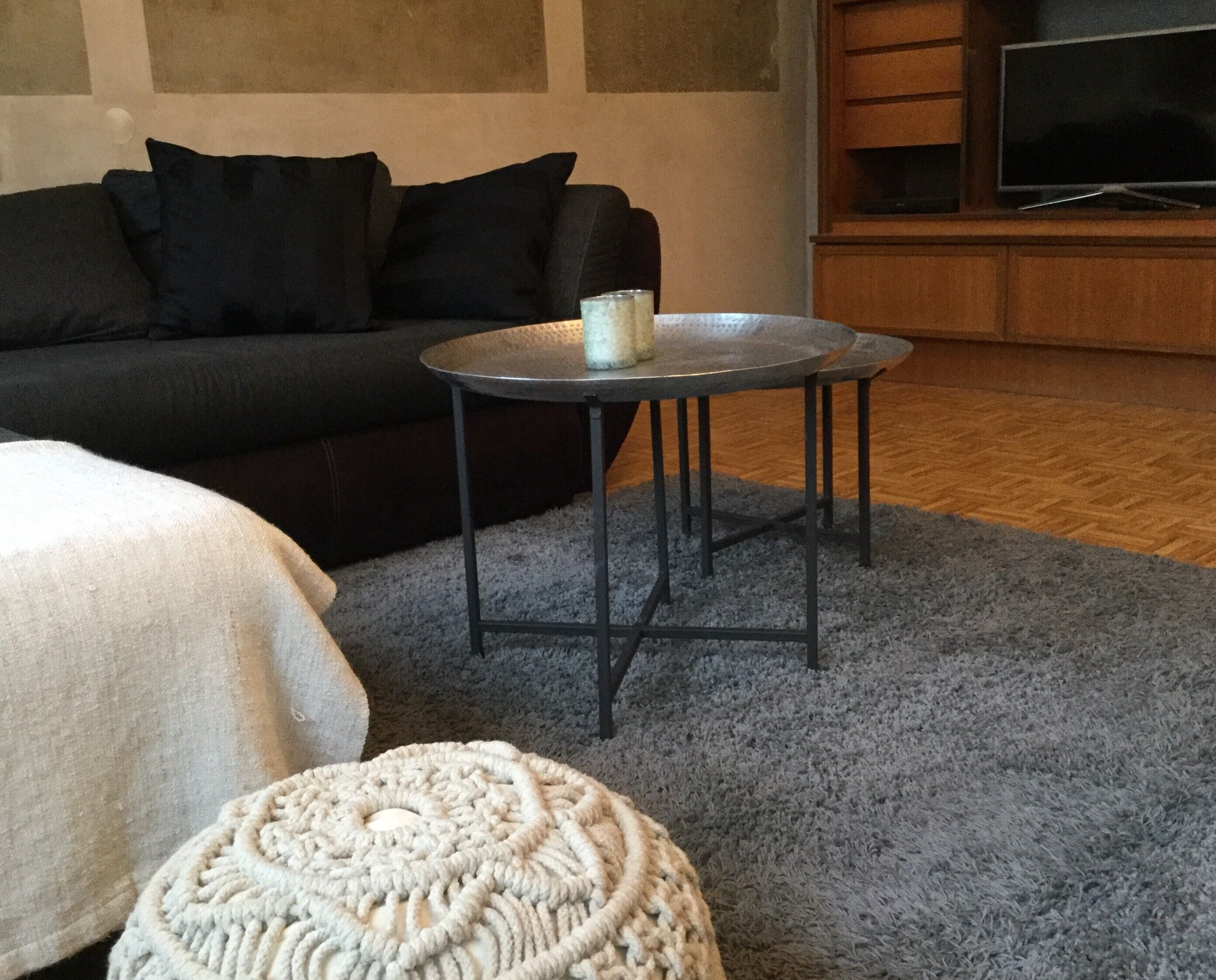 Een woonkamer met een donkere bank, een ronde metalen tafel met een kaars, en een geweven vloerkleed.