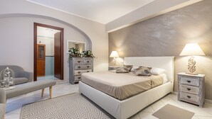 A bedroom with a bed, bedside tables, a bench, a mirror, a dresser, and a door leading to a bathroom. - Apartment "Suite Home Simius" Close to Beach with Terrace, Sea View & Air Conditioning (Villasimius)