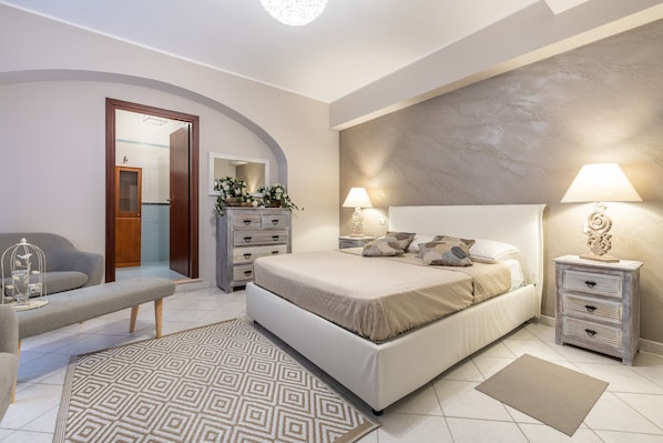 A bedroom with a bed, bedside tables, a bench, a mirror, a dresser, and a door leading to a bathroom.