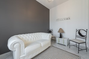 Un canapé en cuir blanc, un tapis à motifs, une lampe, une chaise et une décoration murale en forme d’étoile dans une pièce dont le mur est gris et le sol est recouvert de carreaux blancs.