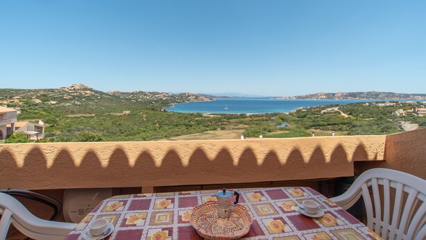 Un balcon avec une table et des chaises, donnant sur un paysage côtier verdoyant et sur une étendue d’eau.