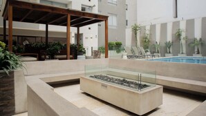 A modern outdoor pool area with a glass-enclosed water feature, lounge seating, and a wooden pergola.