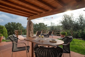 Un patio al aire libre con una mesa y sillas de madera, rodeado de plantas en macetas y vistas a árboles.