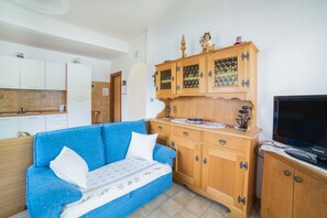 Un salon avec un canapé bleu, des meubles en bois, un téléviseur et, au fond, un coin cuisine.