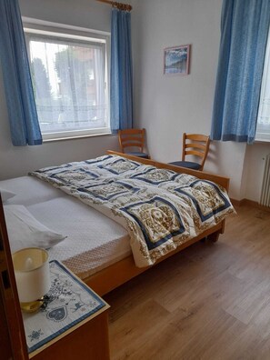 Une chambre à coucher avec un lit, deux chaises, une fenêtre avec des rideaux, un radiateur et un tableau encadré au mur.