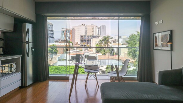Una sala de estar moderna con un ventanal grande, un comedor con mesa y sillas de vidrio, y una cocina con refrigerador y armarios.