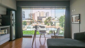 Una sala de estar moderna con un ventanal grande, un comedor con mesa y sillas de vidrio, y una cocina con refrigerador y armarios.