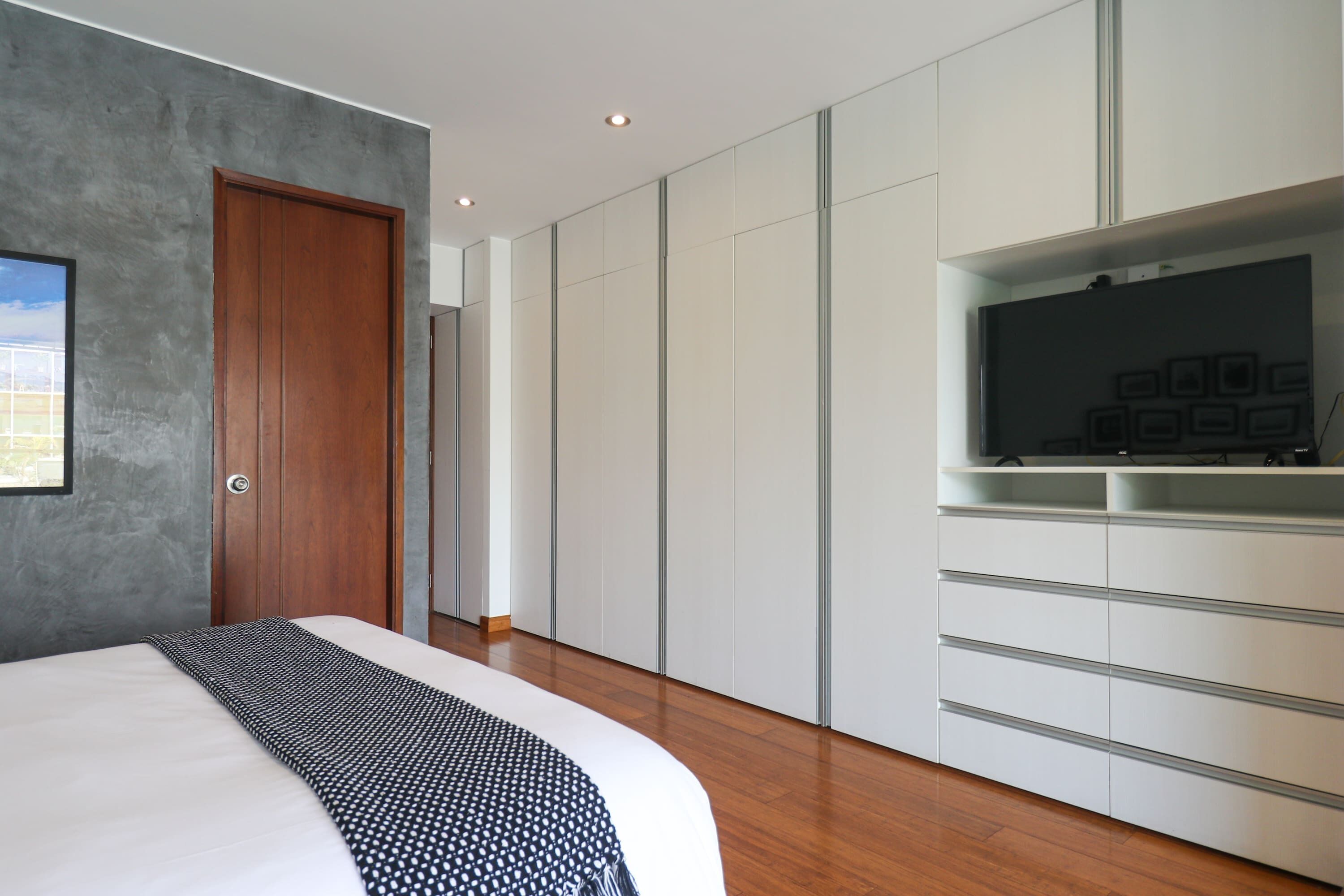 A modern bedroom with a wooden door, a bed with a patterned blanket, a built-in TV shelf, and a wardrobe with sliding doors.