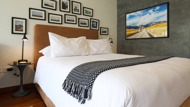 Una cama bien hecha con sábanas blancas y un plaid blanco y negro. Varias fotografías enmarcadas decoran la pared sobre la cama.