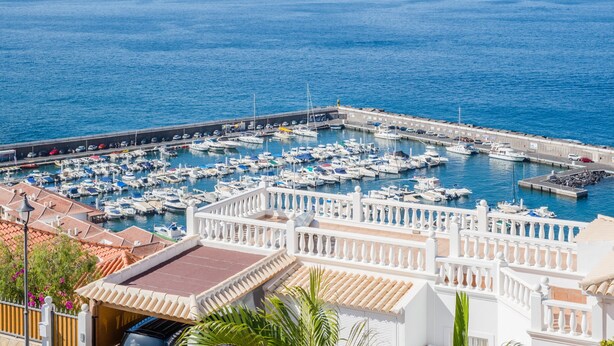 A marina with numerous boats docked, a white waterfront building with balconies, and a clear view of the sea.