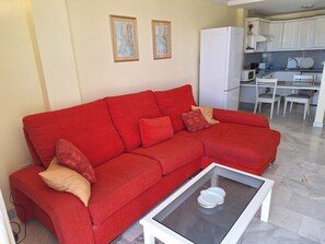 Ein rotes Sofa mit gemusterten Kissen, ein Couchtisch mit Glasplatte und eine Küchenzeile mit weißen Schränken und einem weißen Kühlschrank.