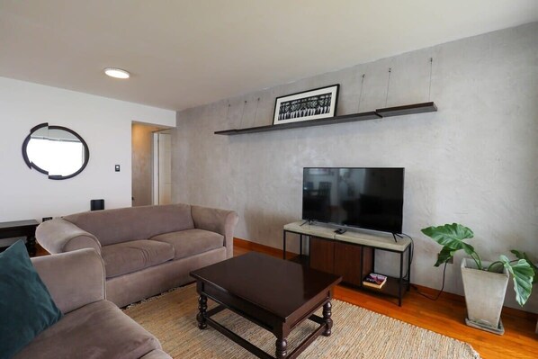 A living room with a sofa, a coffee table, a TV on a stand, a framed picture on the wall, and a potted plant. - w* | Bright 3BR with Ocean View (Lima)