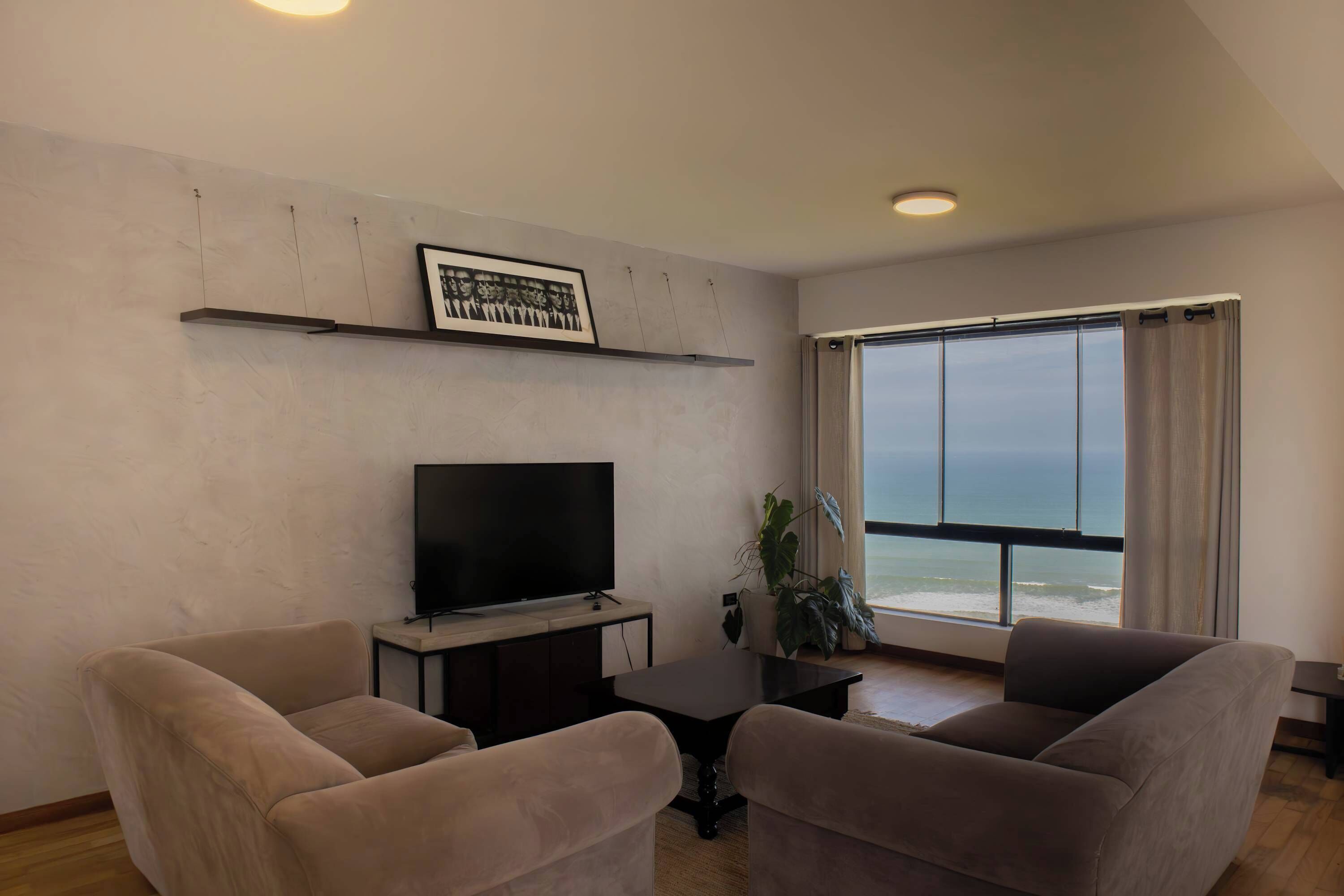 A modern living room with a flat-screen TV, two sofas, a coffee table, and a large window with a view of the ocean.