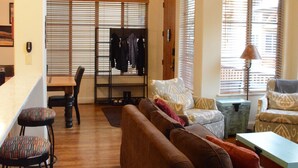 A spacious living area with a wooden floor, a brown sofa, a dining table with chairs, and large windows with blinds. - Lovin' Life. Near all the Fun Stuff! Now with A/C! (Ketchum)