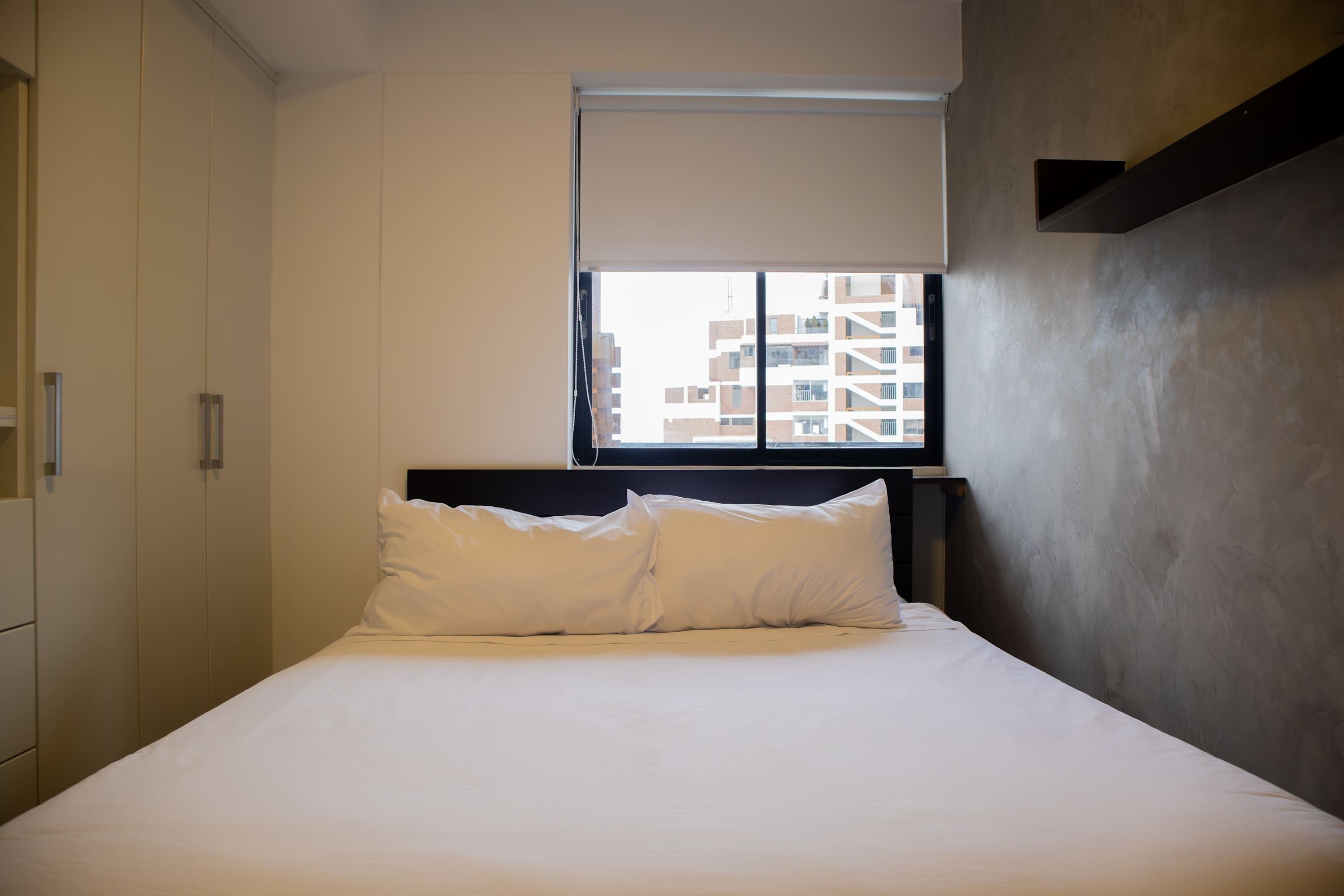 A hotel room with a bed, a window with blinds, a wardrobe, and a view of buildings outside.