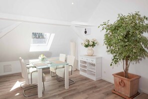 A modern loft bedroom with a glass dining table, white furniture, a large potted plant, and a skylight.