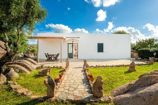 A white building with a stone pathway leading to a door, surrounded by rocks and greenery.
