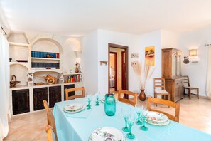 A dining area with a table set for four, surrounded by wooden chairs. There is a bookshelf with books and decorative items, a wooden cabinet, and a door leading to another room.