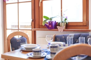 Una mesa de comedor dispuesta con platos, tazas y una copa de vino, con sillas y una ventana al fondo.