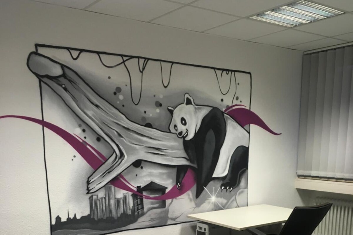 A hotel room with a bed, a desk, and a chair. There is a large wall mural featuring a panda and a cityscape.