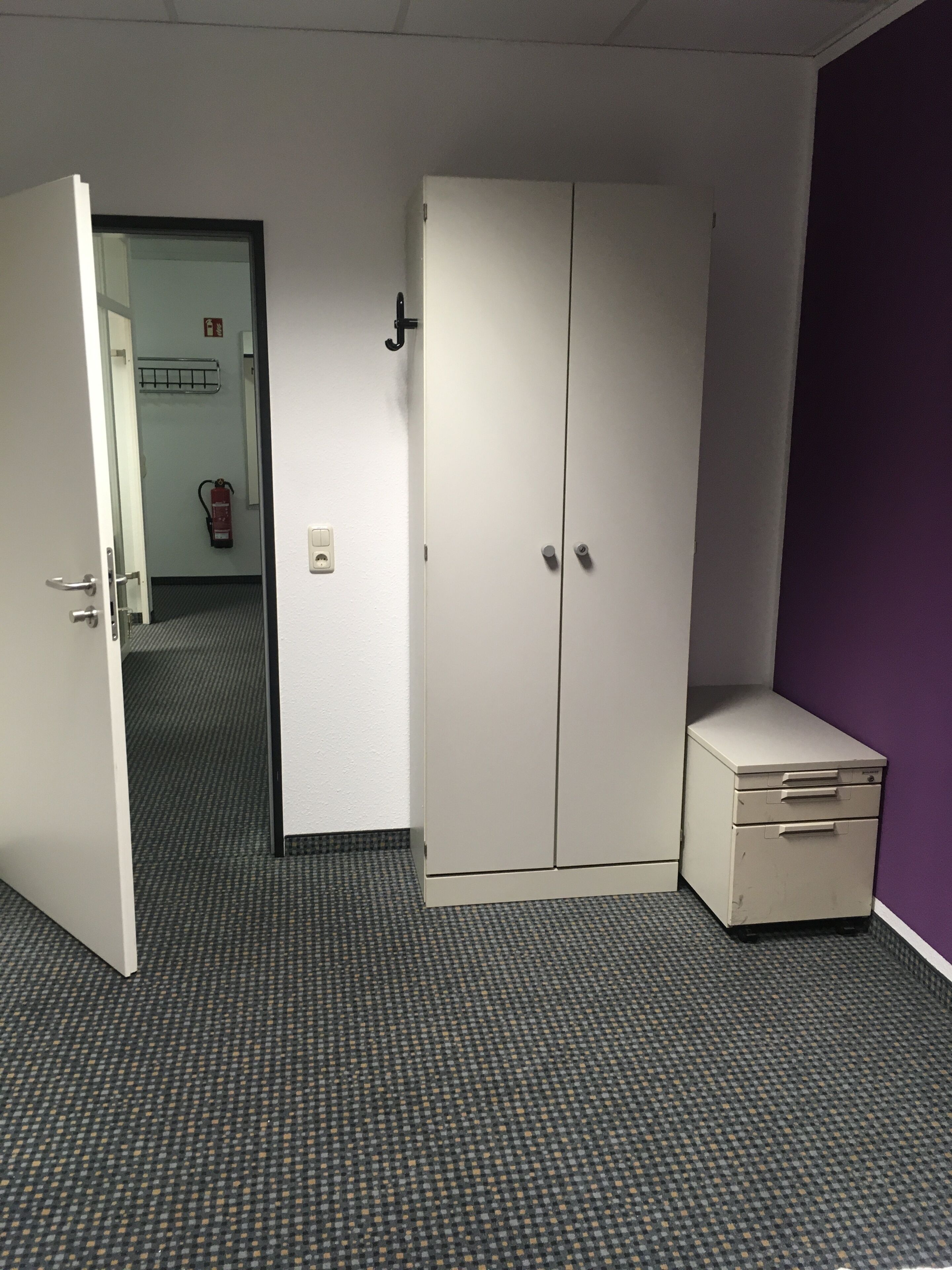 Een kamer met een gesloten deur, een brandblusser, een klein kastje en een kamerbreed tapijt.