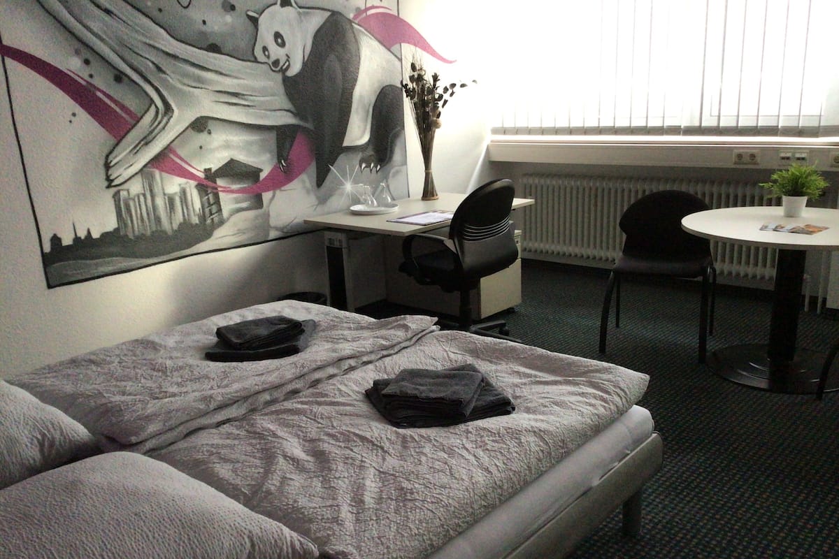A hotel room with a bed, a desk, a chair, a small table, and a radiator.