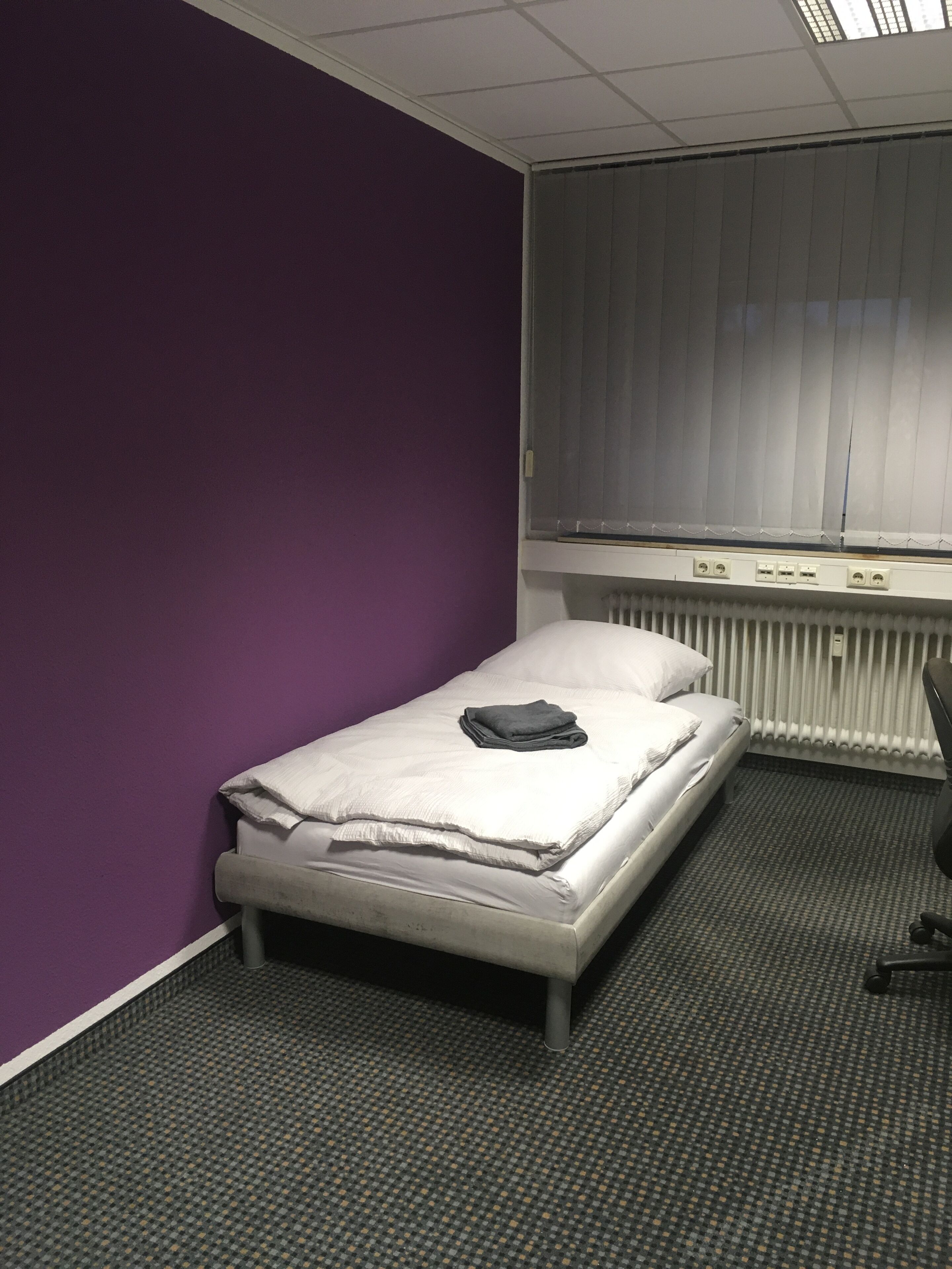 A small, single-bed room with a purple wall, a window with blinds, a radiator, and a desk with a chair.