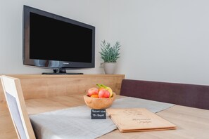 Un salon moderne avec un téléviseur à écran plat, un saladier en bois rempli de fruits, un carnet et une petite plante.