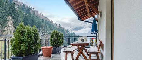 Un balcón con mesa y sillas de madera, plantas en macetas y vista a un paisaje montañoso.