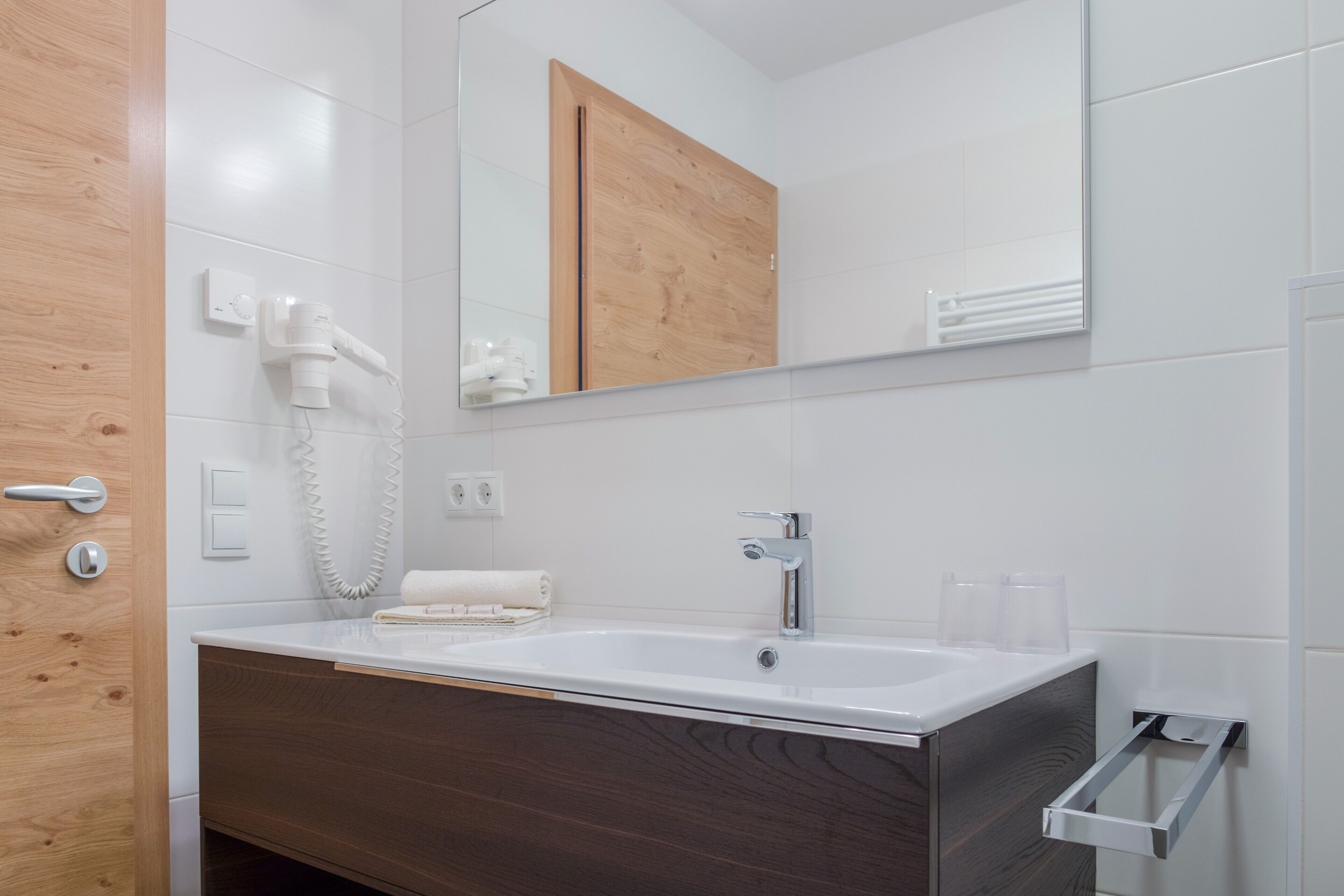 A bathroom with a white sink, wooden vanity, a mirror, a hairdryer, and a towel rack.